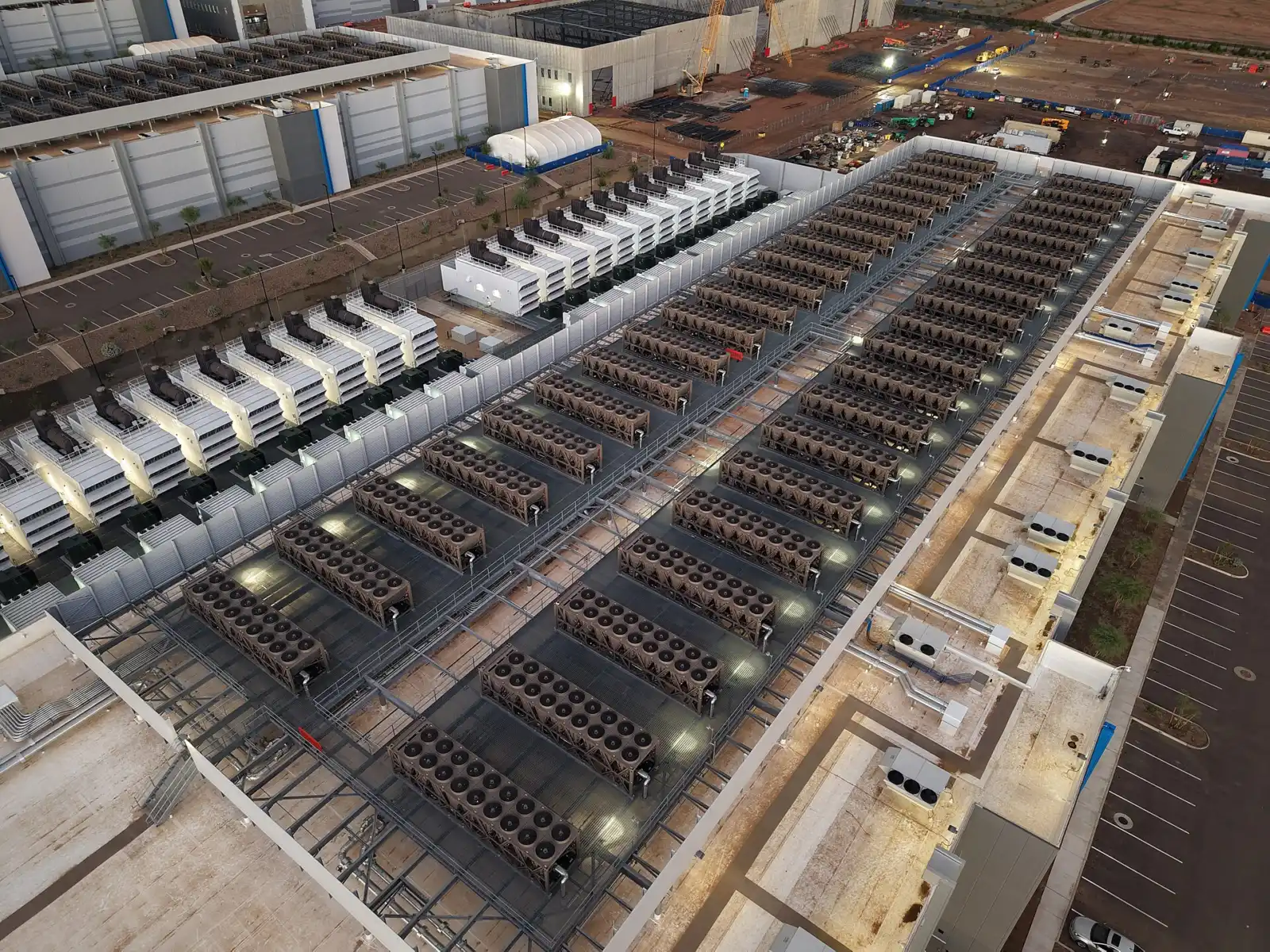 Aerial view of a data center roof lined with rows of fan-topped cooling units, beside a row of backup generator enclosures and a campus still under construction